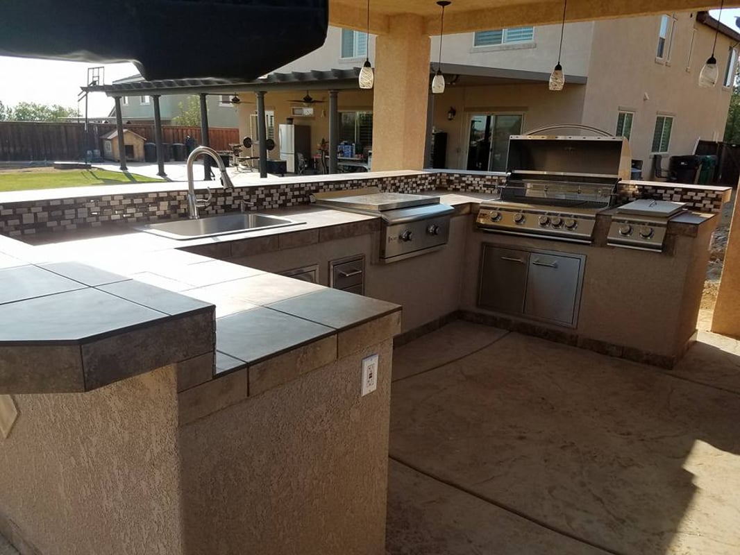 Covered U-shaped outdoor kitchen with sink, grill, side burner, and tiled backsplash